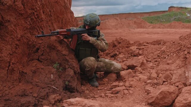 Camouflaged soldier kneels in red clay ravine aiming rifle with focused posture using earthen cover during military training outdoors under cloudy sky side view