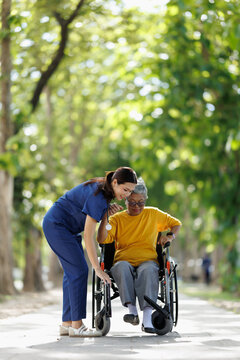 Young nurse in scrubs providing thoughtful support to an elderly asian woman sitting in a wheelchair, moving along a shaded pathway outdoors, focusing on caregiving and assistance