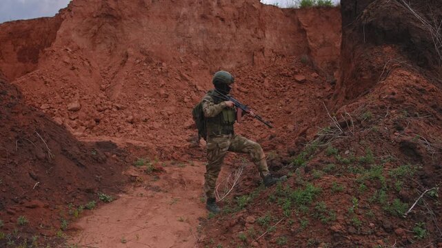 Soldier in tactical gear aiming rifle while advancing across steep red clay slope, alert stance on rugged desert terrain with embankment and overcast light, with soldier, rifle, aiming, tactical