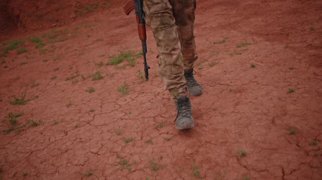 Lower body of armed soldier walking across cracked red earth, muddy camouflage trousers and worn combat boots moving through sparse green sprouts in harsh rural terrain with tense patrol mood