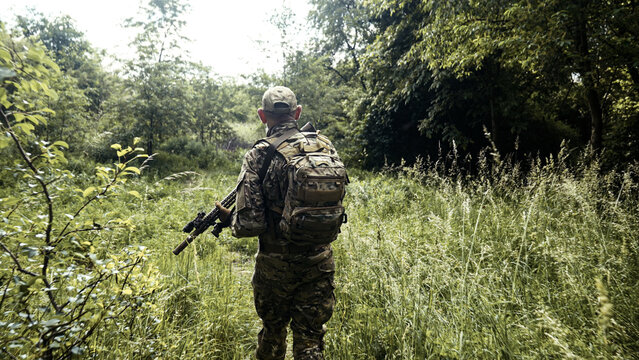 Camouflaged soldier in the middle of a field of wild grass on a mission. Walking and aiming a AR-15 carbine rifle with a red laser collimator and a magnifier