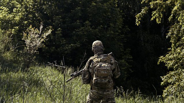 Camouflaged soldier in the middle of a dark forest on a recon mission. Walking and aiming a AR-15 carbine rifle with a red collimator and a magnifier
