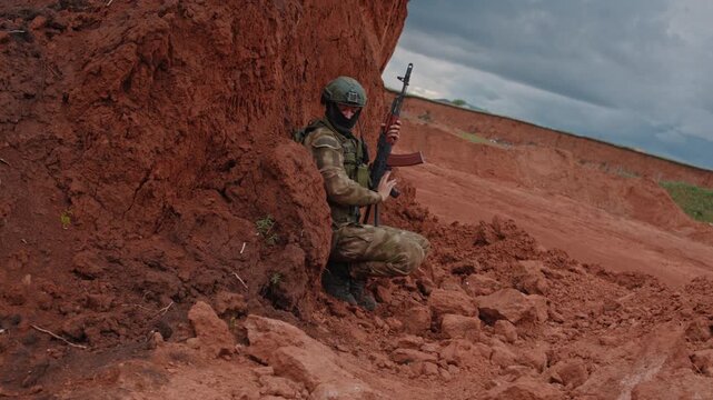 Camouflaged soldier crouches beside red clay embankment, holding rifle upright while facing camera in rugged rocky terrain under cloudy sky, with tense defensive stance and stark outdoor detail