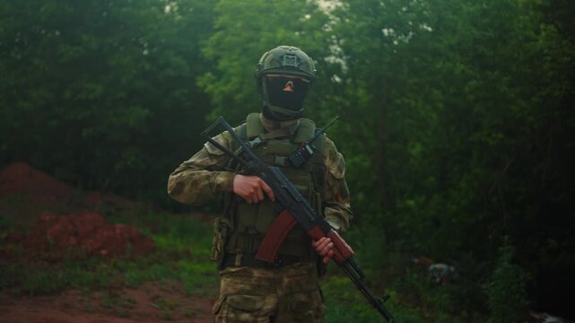 Camouflaged soldier with rifle stands alert on forest path, surrounded by dense green trees, side view, slightly low angle, tactical gear, backpack, helmet, radio, and watchful military stance