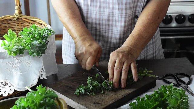 Close-up of senior woman hands cutting fresh organic green kale vegetables on a wooden chopping board, preparing a healthy homemade superfood meal in the kitchen.