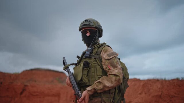 Soldier lowering rifle while adjusting sling on red earth training ground beneath overcast sky, full tactical gear, anonymous subject, outdoor military practice