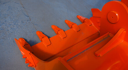 The teeth are sharp and new. The excavator bucket is used to move soil using a mechanical arm. The equipment is painted orange on a summer day. © Elena