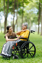 Obraz premium Young asian caregiver providing compassionate assistance to a senior woman in a wheelchair, listening to her concerns outdoors in a peaceful park setting