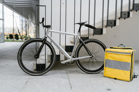 White Fixed-Gear Bicycle With Yellow Delivery Backpack Parked Outside Modern City Building