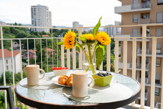Glass table on balcony decorated with sunflowers and served breakfast croissants, coffee mugs and berries. Cozy outdoor urban lifestyle