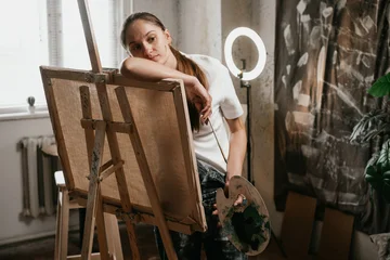 Fotobehang Schilderij Depressed woman painter standing near easel with palette in hands in creative workshop. Concept of artistic crisis, emotional exhaustion, sadness, loneliness and burnout.  © Andrii Lysenko