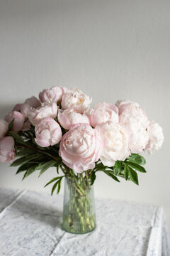 Vertical shot of lush pink peony bouquet with green leaves in ribbed glass vase on table against neutral grey wall in natural light.
