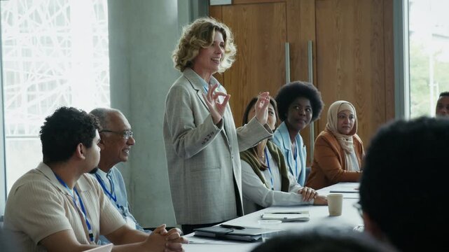 A confident woman presents to an attentive group of professionals gathered at a conference. They engage in discussion, actively listening and contributing to foster collaboration and understanding.