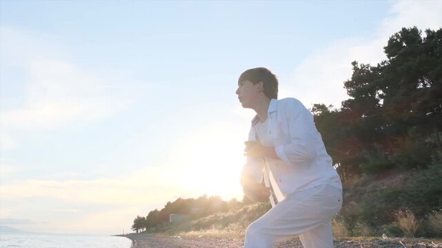 Handsome boy with beautiful hairstyle wearing white clothes throwing stones into sea on seashore at sunset while alone. Young man alone with his thoughts in nature. Concept of solitude with yourself