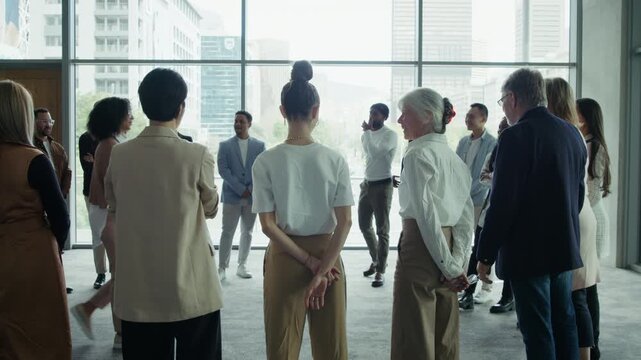 Business professionals in a spacious room with city views. They gather in a circle, conversing and networking at a corporate event, fostering connections and collaborative spirit.