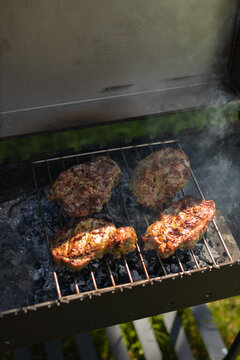 High angle shot of meat pieces grilling on metal rack over hot charcoal with smoke in outdoor barbecue grill. Ideal for summer food and restaurant advertising