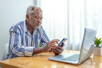 Fototapeta premium Mature man with white beard casually using phone and laptop at home.
