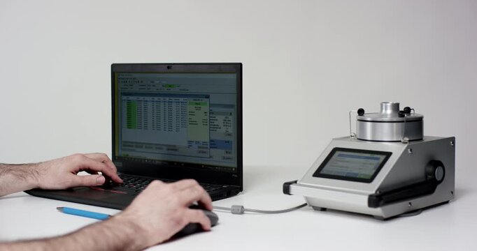 A person types on a laptop connected to an electronic laboratory device on a white surface for data collection.