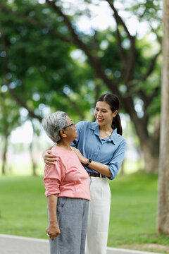 Young asian woman supporting a senior woman walking outdoors, showing care, companionship, and a strong intergenerational relationship in a green park setting