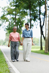Fototapeta premium Senior asian woman and younger relative walking hand in hand in a green park, enjoying a healthy outdoor stroll and showing intergenerational support