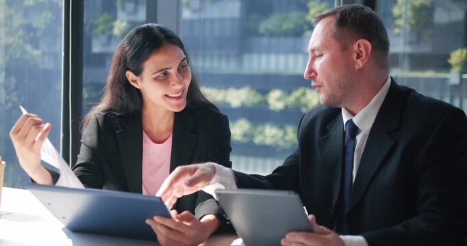 Business Collaboration: A man and woman in business attire engage in a lively discussion over documents and tablets, showcasing teamwork and effective communication in a modern office setting.