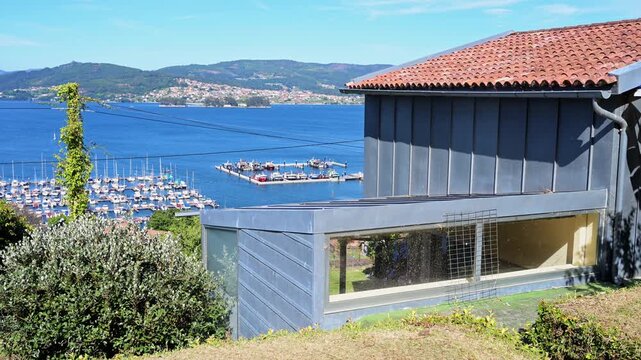 Summer panorama of a hillside residence with zinc roofing facing the Vigo estuary boat dock pier in Galicia, Spain. Documents premium coastal real estate development and nautical travel vistas.