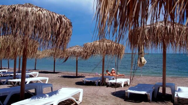 Tropical beach resort scene with thatched straw umbrellas, white sun loungers, and two girlsrelaxing on the sandy shore near the sea. 