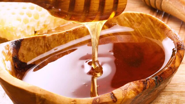 Close up shot of a wooden honey dipper drizzling sweet organic golden honey into a bowl against a warm rustic kitchen background