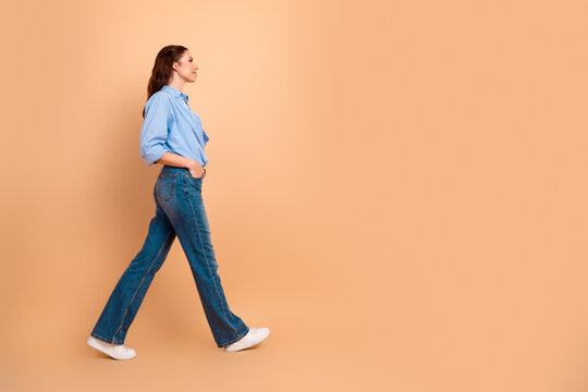 Woman walking in profile across a peach background wearing blue shirt and jeans, casual lifestyle and confident stride in studio
