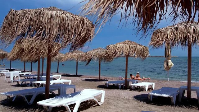 Tropical beach resort scene with thatched straw umbrellas, white sun loungers, and two girlsrelaxing on the sandy shore near the sea. 