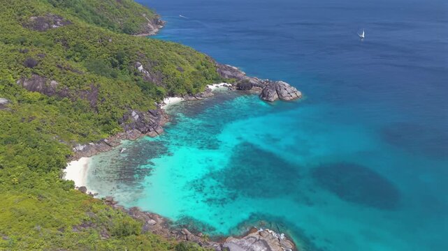 Rotating drone shot of the secluded Anse Du Riz cove in Mahe, Seychelles, featuring a luxury catamaran sailing in turquoise marine park waters below lush granite mountains.