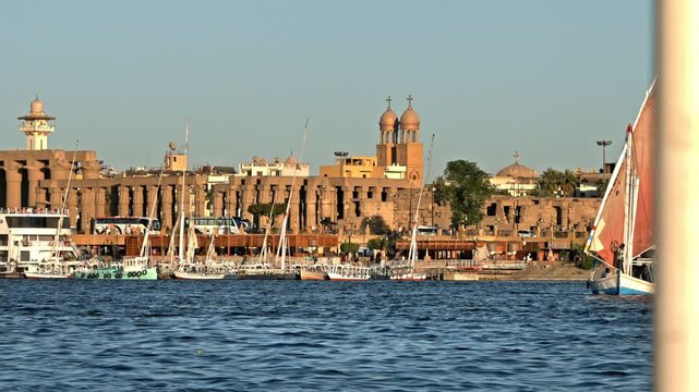 A wide sunset view looking across the blue waters of the Nile River toward the Luxor riverbank, lined with docked felucca sailboats, the ancient columns of Luxor Temple, and prominent Christian chur