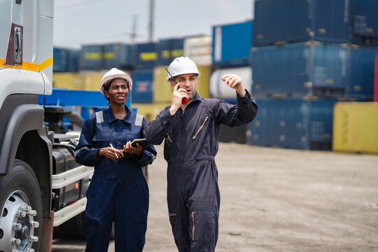 Container yard supervisor speaking on two-way radio while discussing cargo operations with logistics colleague beside a freight truck. Global shipping, container terminal, transportation planning.