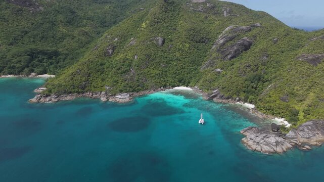 Aerial drone footage of a luxury catamaran anchored in the vibrant turquoise waters of Anse Du Riz cove, surrounded by lush tropical mountains and granite rocks in Mahe, Seychelles