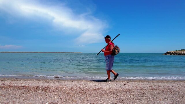 Fisher man walking on a sunny beach holding a fishing rod over his shoulder, walking past a seagull near the sea shore. 