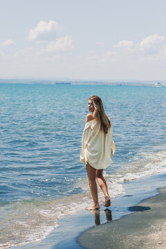 Woman walks along the shore by the blue ocean in a sunny location during the day with a light breeze and clouds in the sky