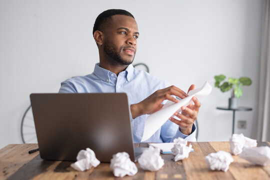Young black man working in an office. Facing writer's block while trying to complete a task. Sitting at a desk with a laptop and many crumpled papers
