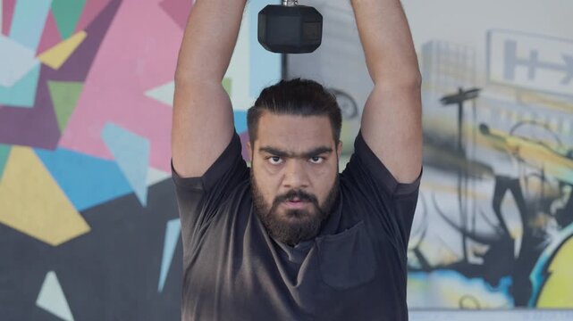 Bearded male athlete performing a standing overhead dumbbell triceps extension during a strength training session in a modern indoor fitness studio.
