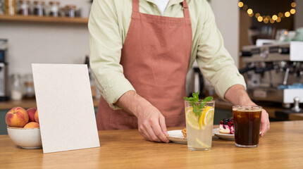Fototapeta premium Barista serving lemonade, iced coffee and small desserts on a wooden cafe counter, with a blank menu card for copy space and a warm coffee shop interior
