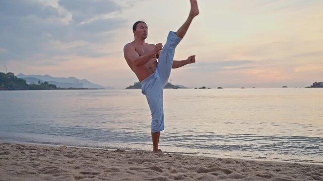 Shirtless martial artist training on sandy beach at sunrise preparing low walking kick during middle of coastal sequence with side light and distant islands. slow motion