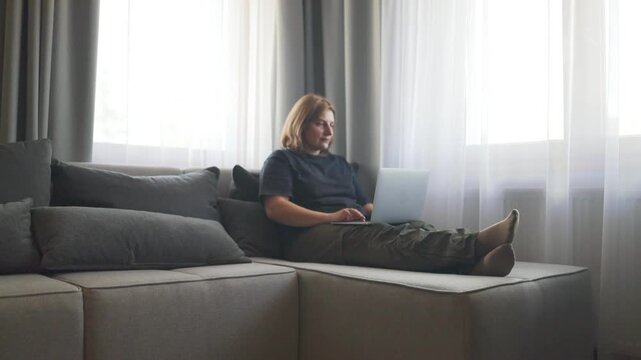 Woman sitting and using a laptop, in modern living room with large window, with laptop, couch, with textile couch glass window texture, from a side view perspective, with gray, white accents.