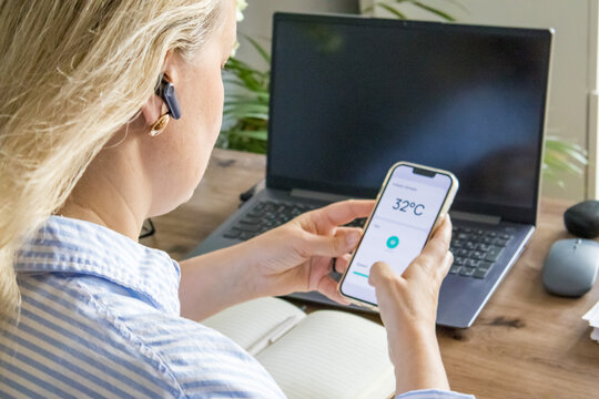 Close-up of a smartphone displaying an indoor temperature of 32 degrees Celsius while standing beside a laptop in a home office. Smart temperature monitoring, heatwave and connected home technology.