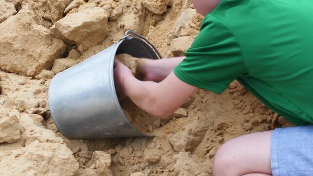 Detail of a little kid's hands playing with yellow sand and a metal pail in a sandbox or on a construction site, enjoying a sunny day outdoors during childhood summer vacation