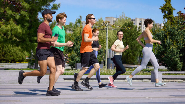 Young fitness group jogging together through green city park. Concept of cardio training, running club, active lifestyle, wellness, friendship and outdoor sport.