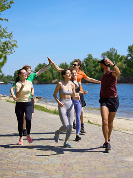 Running friends celebrate with high five on sunny riverside path. Concept of fitness motivation, team support, active lifestyle, outdoor cardio and wellness community.