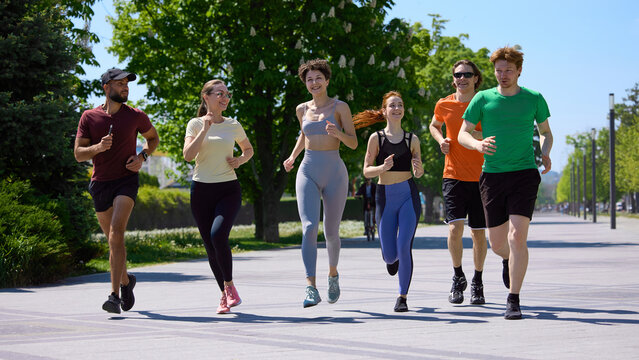 Group of young runners jogging together on sunny park promenade. Concept of running club, group fitness, healthy lifestyle, urban recreation and outdoor cardio training.