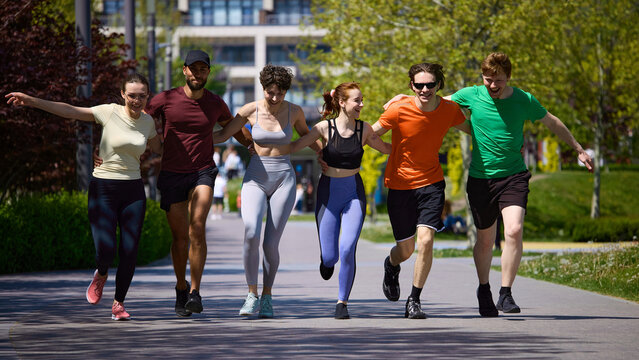 Friends running together with arms around shoulders in sunny park. Concept of teamwork, motivation, fitness friendship, running club culture, healthy lifestyle and outdoor exercise.