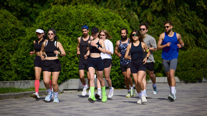 Large running club jogging together on sunny park path. Concept of marathon training, group cardio...