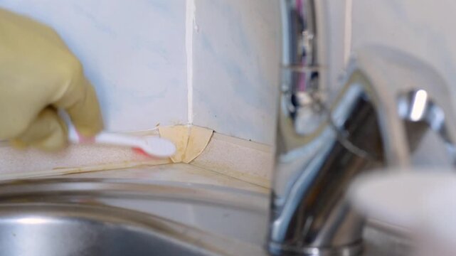 Gloved hand applying cleaning paste to corner grout by faucet, closeup of precision toothbrush work on tile joint, focus on removing brown buildup and preparing for scrubbing,
