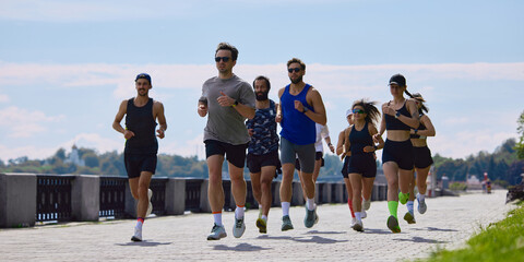 Mixed running club jogging on sunny riverside path with city skyline. Concept of marathon preparation, group fitness, wellness programs and active weekend lifestyle. © master1305